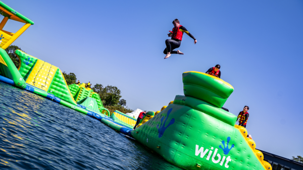 People enjoying an inflatable water park on a sunny day, with one person jumping into the water from a large green structure while others watch.